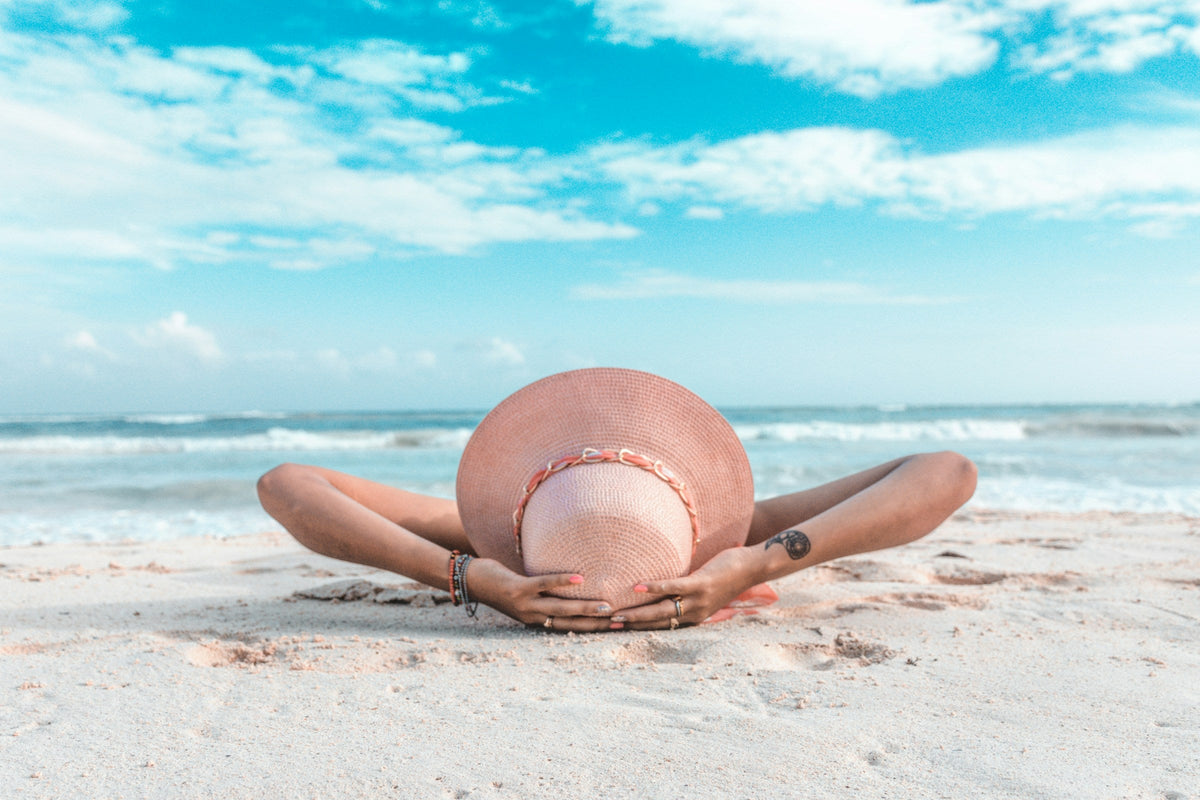 Flat lay of essential beach vacation items including swimwear, towel, sunscreen, and a straw hat on white sand