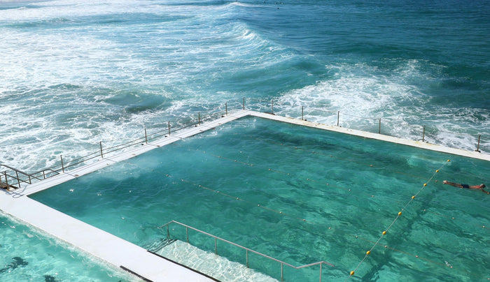 Aerial view of a dramatic cliffside infinity pool overlooking the ocean, with swimmers enjoying a panoramic sunset