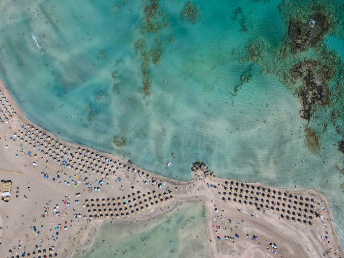 Aerial view of a family-friendly beach in Europe with shallow turquoise water, soft white sand, children playing, and colorful umbrellas