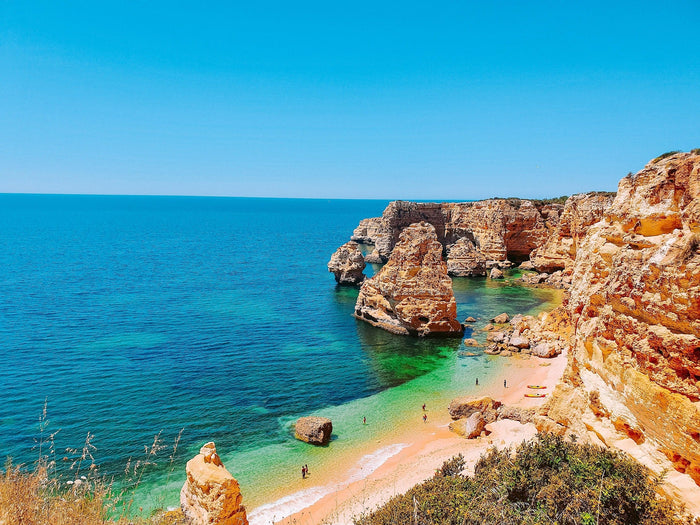 Aerial view of a European beach with turquoise water, golden sand, and dramatic rocky cliffs — perfect for a slow and stylish summer